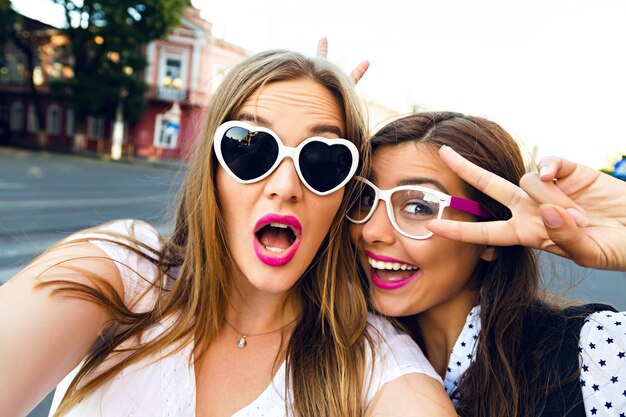 Летний солнечный образ двух лучших подруг сестер, брюнетки и блондинки, веселых на улице, делающих селфи, в забавных винтажных солнцезащитных очках, яркого стильного макияжа с длинными волосами