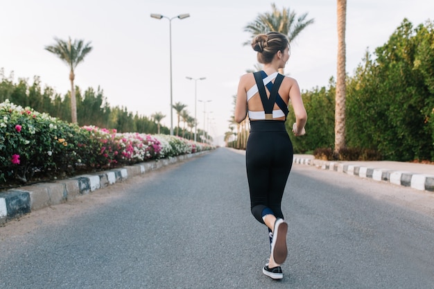 Ora legale di gioiosa giovane donna dal retro in esecuzione su strada con palme, fiori colorati in città tropicale. stato d'animo allegro, divertimento, allenamento, mattina di sole, modello attraente.