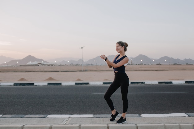 Ora legale di gioiosa giovane donna in abiti sportivi a piedi, guardando l'orologio a portata di mano, sorridente sulla strada nel paese tropicale. stato d'animo allegro, allenamento, mattina di sole, modello attraente.