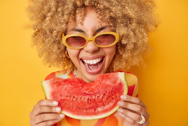 여름 분위기 긍정적인 곱슬머리 유럽 여성은 맛있는 수박 조각을 먹고 노란색 배경에 격리된 트렌디한 선글라스를 끼고 큰 조각을 들고 건강한 맛있는 음식 개념