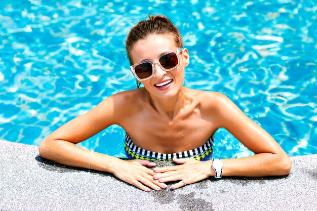 Summer trendy close up fashion portrait of sexy tanned woman relaxed and swimming at pool. wearing bright bikini and sunglasses, smiling and looking