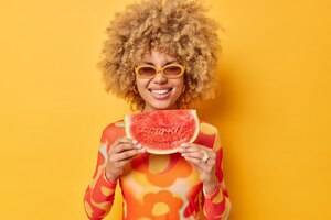 summer vibes concept positive curly haired beautiful woman holds big slice of juicy red watermelon feels very happy wears sunglasses and casual jumper isolated over vivid yellow background
