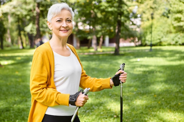 Summertime, active lifestyle, leisure and hobby concept. Outdoor shot of healthy energetic elderly female in yellow cardigan walking in park on sunny day using Nordic poles, having happy look