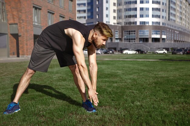 Summertime, activity, fitness and hobby concept. Muscular young European unshaven man in black sports clothes and running shoes bending over to left toe, stretching legs while exercising outdoors