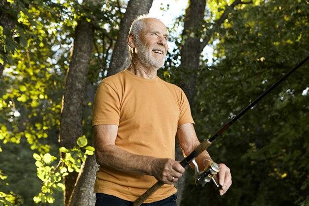 Summertime picture of handsome energetic active unshaven senior man on retirement spending nice summer morning outdoors, catching fish using fishery rod, having joyful happy facial expression