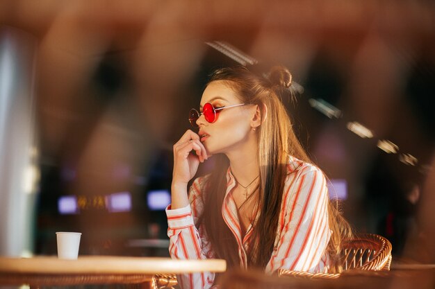 Sun shines over the faces of stunning woman sitting in red sunglasses in the cafe