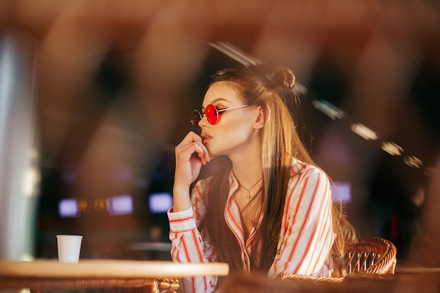 Sun shines over the faces of stunning woman sitting in red sunglasses in the cafe
