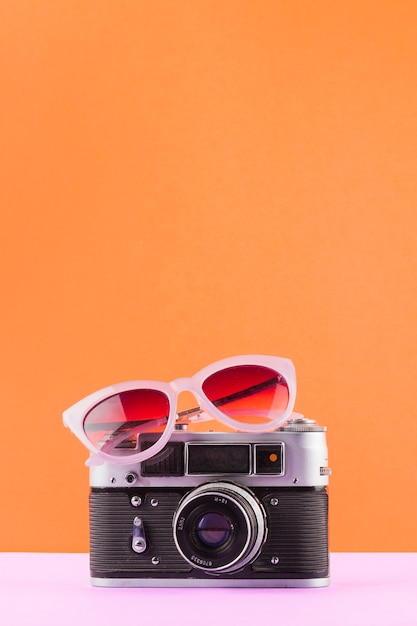 Sunglasses over the vintage camera on white desk against an orange background