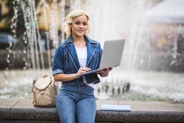 日当たりの良いブロンドの女の子の女性学生は日に市内の噴水の近くの彼女のラップトップコンピューターで動作します