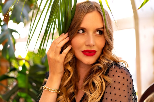 Sunny close up portrait of pretty young curly woman posing near tropical leaves winter garden, evening glamour outfit and make up.