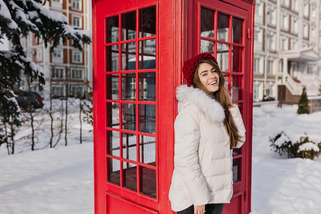 Mattinata soleggiata congelata di una giovane donna alla moda che sorride sulla strada invernale piena di neve. incredibile ragazza in abiti invernali caldi che si gode il freddo sullo sfondo della cabina telefonica rossa