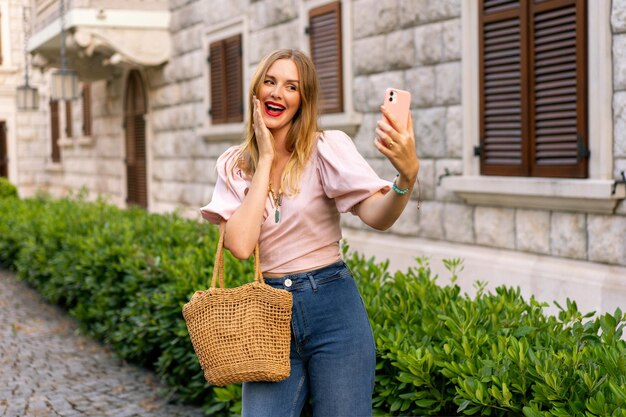 Sunny outdoor image of stylish blomde woman travel in europe spring summer vacation 70s outfit making selfie on the street holding smartphone