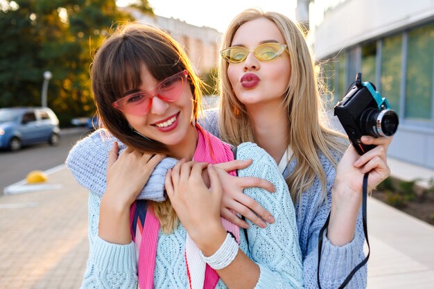 日当たりの良い屋外のポートレートまたは2つの陽気な面白い流行に敏感な女性パステルカラーの流行のセーターとメガネを着てビンテージカメラでselfieを作る、姉妹の親友が一緒に楽しんで。