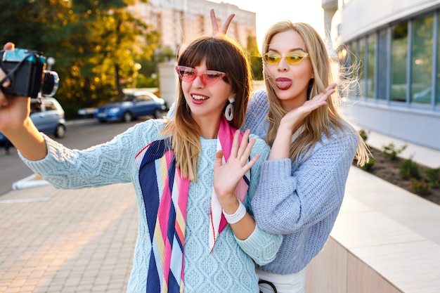 Sunny outdoor portrait or two cheerful funny hipster woman making selfie on vintage camera wearing pastel colors trendy sweaters and glasses, sister best friends having fun together.