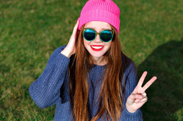 Sunny positive portrait of happy smiling hipster woman, posing at park, travel, vacation, joy, shows v  by her finders, spring mood, sweater and hat.