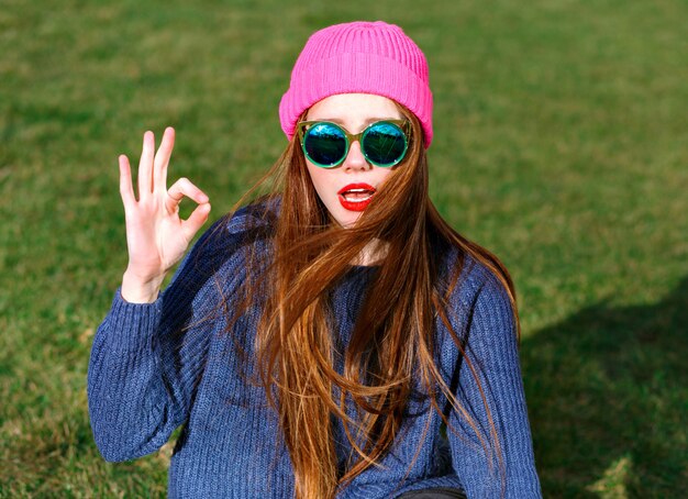 Sunny positive portrait of happy smiling hipster woman, posing at park, travel, vacation, joy, shows v  by her finders, spring mood, sweater and hat.