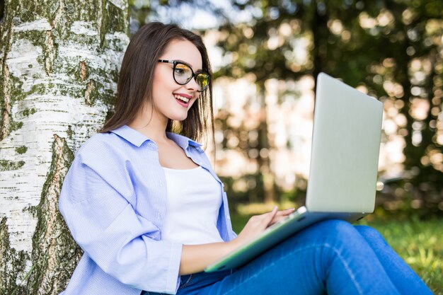 Солнечная красивая девушка в синих джинсах работает с ноутбуком в ситипарке