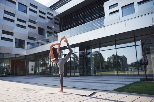 Sunny summer morning. Young athletic woman doing handstand on city park street among modern urban buildings. Exercise outdoors healthy lifestyle