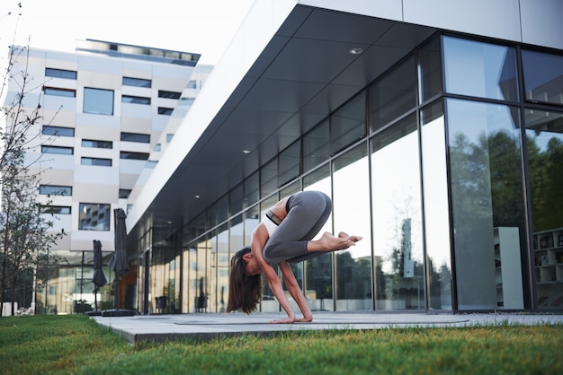 Sunny summer morning. Young athletic woman doing handstand on city park street among modern urban buildings. Exercise outdoors healthy lifestyle