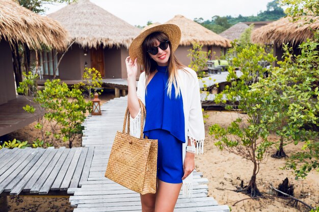 Солнечный летний портрет милой брюнетки в соломенной шляпе и модной сумке бохо, идущей в тропической вилле в Таиланде.