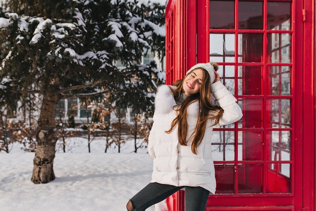 무료 사진 맑은 겨울 아침, 거리에 빨간 전화 박스 근처에서 즐기는 따뜻한 옷을 입은 매력적인 여성의 좋은 분위기. 추운 날씨, 따뜻하고 밝은 감정, 만찬