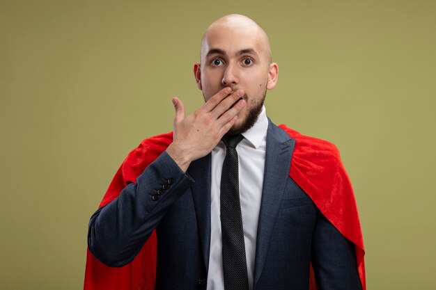 Super hero bearded business man in red cape  covering mouth with hand being shocked standing over light green wall