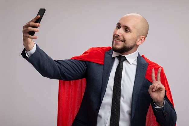 Super hero bearded business man in red cape doing selfie using smartphone smiling showing v-sign standing over white wall