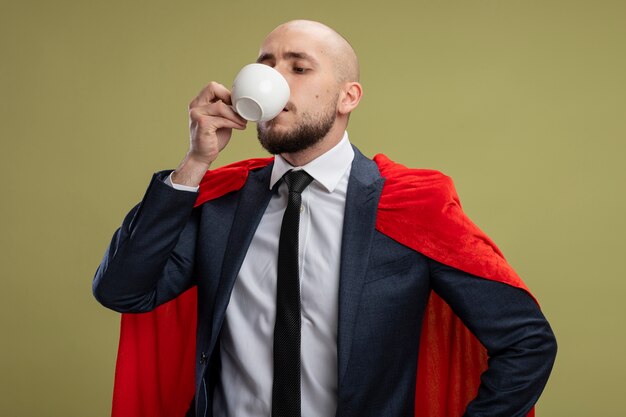 Бородатый супергерой деловой человек в красной накидке пьет кофе и выглядит уверенно, стоя над светло-зеленой стеной