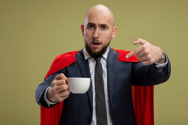 Uomo d'affari barbuto super eroe in mantello rosso che tiene tazza di caffè che punta con il dito indice a essere confuso in piedi sopra la parete verde chiaro