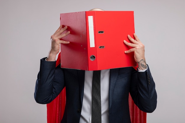 Super hero bearded business man in red cape holding open folder covering his face behind it standing over white wall