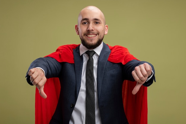 Foto gratuita uomo d'affari barbuto super eroe in mantello rosso che sembra sorridente che mostra i pollici giù in piedi sopra la parete verde chiaro