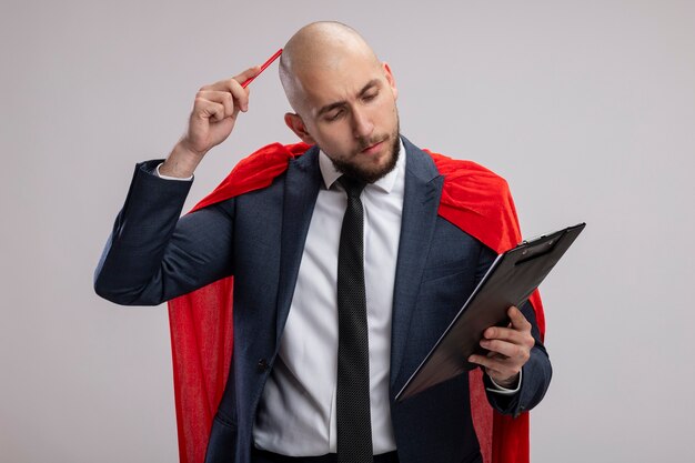 흰색 벽 위에 서있는 펜으로 그의 머리를 긁적 페이지를보고 빈 페이지와 클립 보드를 보여주는 빨간 케이프에서 슈퍼 영웅 수염 비즈니스 남자