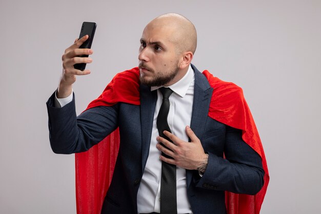 Super hero bearded business man in red cape using smartphone looking at it with serious face worried standing over white wall