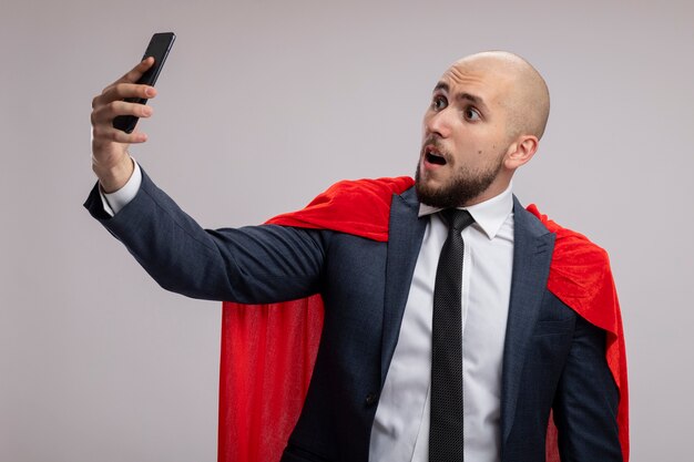 Super hero bearded business man in red cape using smartphonedoing selfie being confused standing over white wall