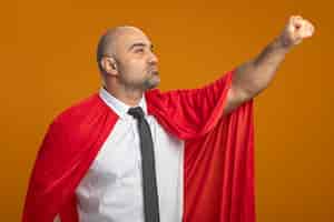 Бесплатное фото Бизнесмен супергероя в красном плаще смотрит в сторону, делая выигрышный жест рукой, готовой помочь выглядеть уверенно, стоя над оранжевой стеной