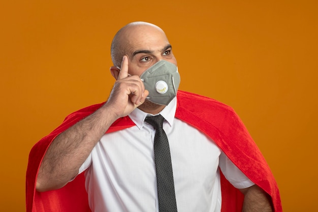 Super hero businessman in protective facial mask and red cape looking aside with pensive expression on face thinking 