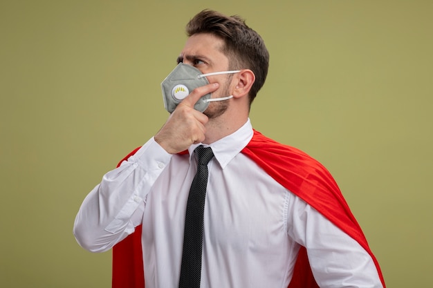 Super hero businessman in protective facial mask and red cape looking aside with pensive expression on face with hand on chin thinking standing over green wall