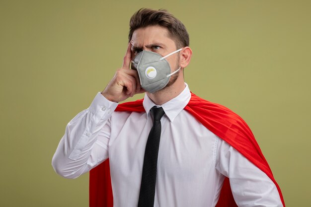 Super hero businessman in protective facial mask and red cape looking aside with pensive expression on face with hand on face thinking standing over green background