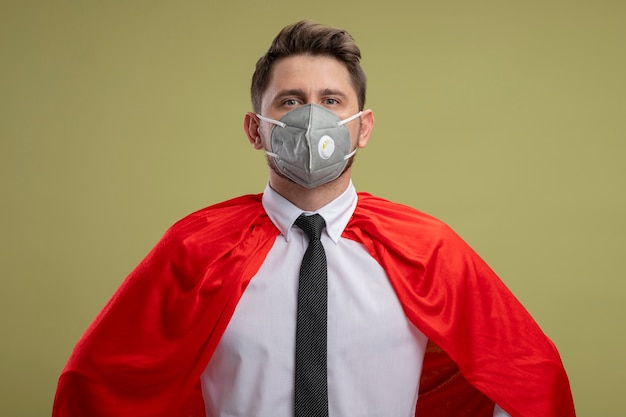 Super hero businessman in protective facial mask and red cape looking confident with arms at hip standing over green wall