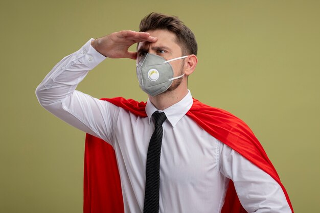 Super hero businessman in protective facial mask and red cape looking far away with hand over head standing over green wall