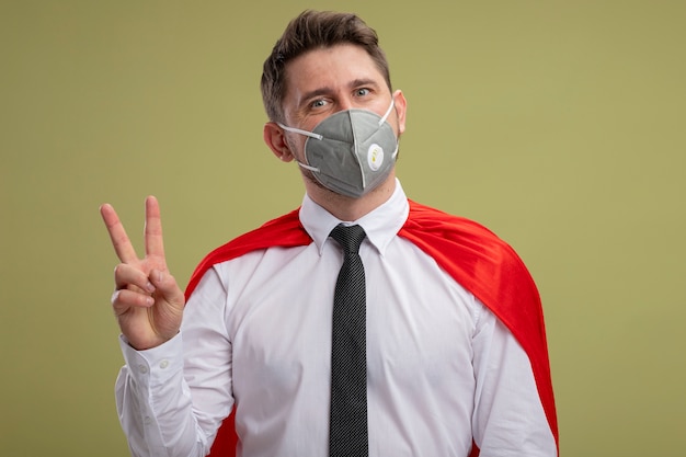 Super hero businessman in protective facial mask and red cape  smiling happy and positive showing v- sign standing over green wall