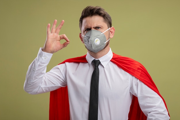 Free photo super hero businessman in protective facial mask and red cape  smiling showing ok sign standing over green wall