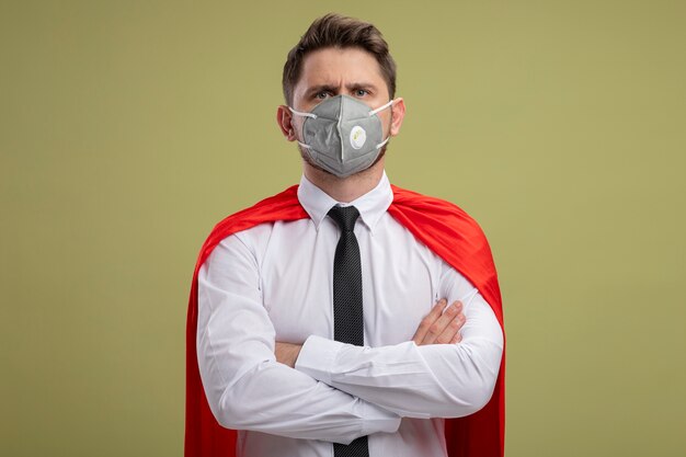 Free photo super hero businessman in protective facial mask and red cape  with crossed hands on chest with serious frowning face standing over green wall