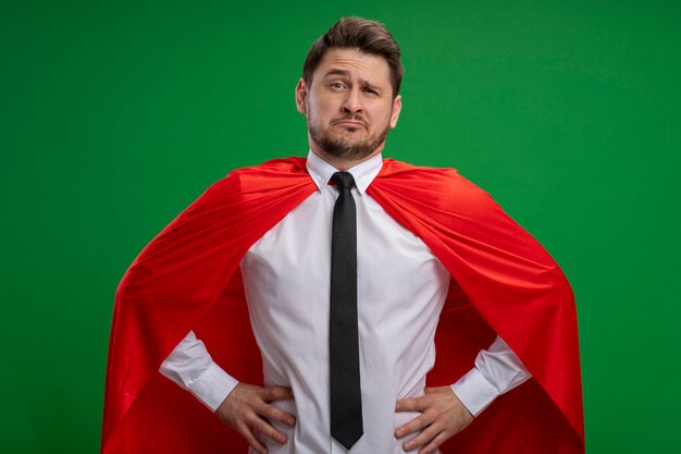 緑の壁の上に立っているヒップで腕と混同されている赤いマントのスーパーヒーローの実業家