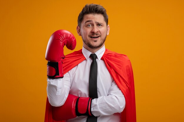 Super hero businessman in red cape and in boxing gloves  confused with raised hand standing over orange wall