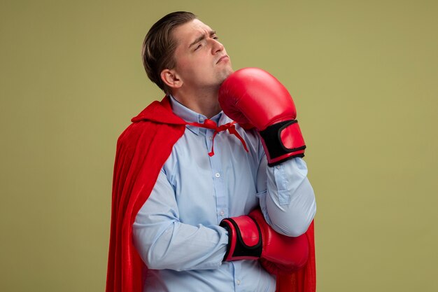 Super hero businessman in red cape and in boxing gloves looking aside with pensive expression on face with hand on his chin standing over light wall