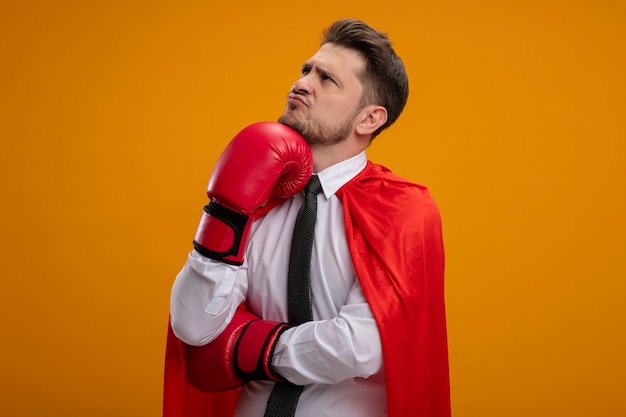 Super hero businessman in red cape and in boxing gloves looking aside with pensive expression on face with hand on his chin standing over orange wall