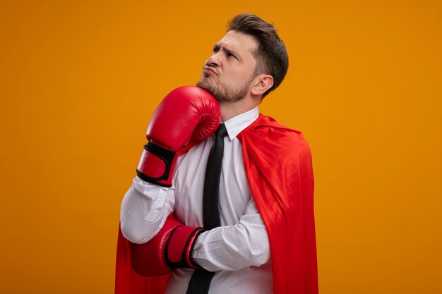 Super hero businessman in red cape and in boxing gloves looking aside with pensive expression on face with hand on his chin standing over orange wall