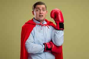 Free photo super hero businessman in red cape and in boxing gloves looking at camera confused with raised hand standing over light background