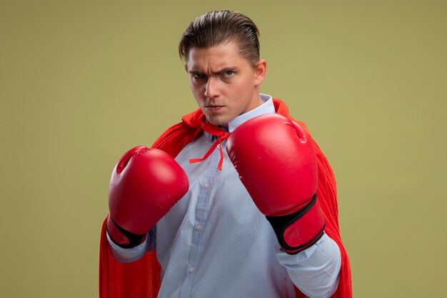 Super hero businessman in red cape and in boxing gloves looking at camera with serious face ready to fight standing over light background